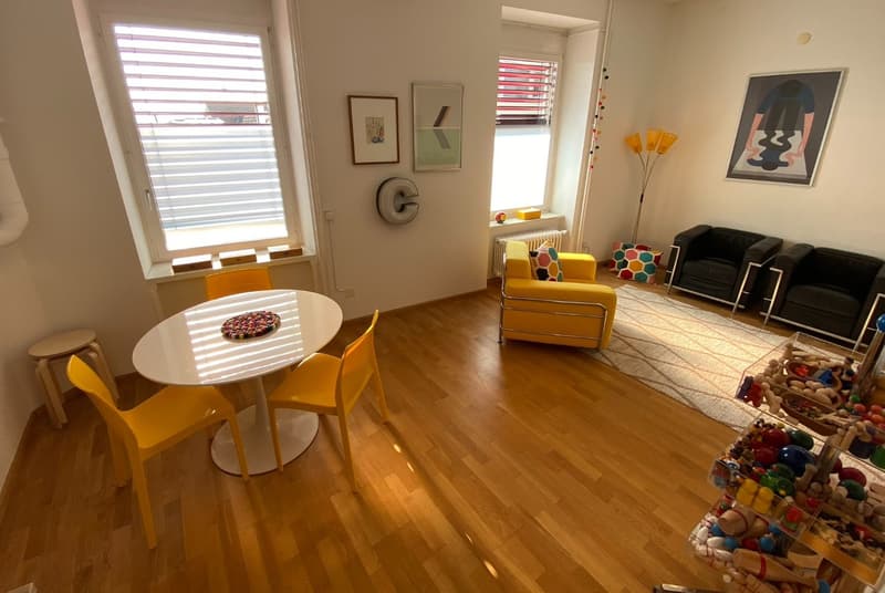 Bright and modern living room with hardwood floors, large windows, a round dining table with yellow chairs, a yellow sofa, and colorful artwork on the walls. The room also has a toy storage area with various toys and games.