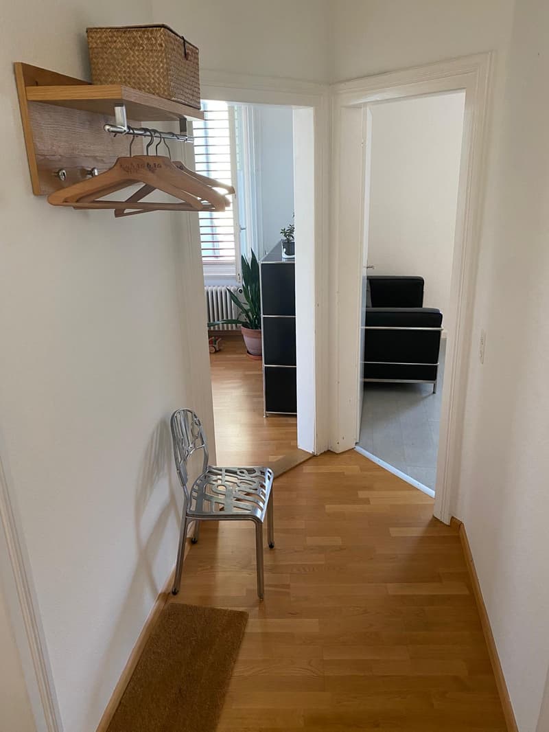 The image shows a bedroom with a wooden closet system, a chair, and a plant. The room has hardwood floors and white walls.