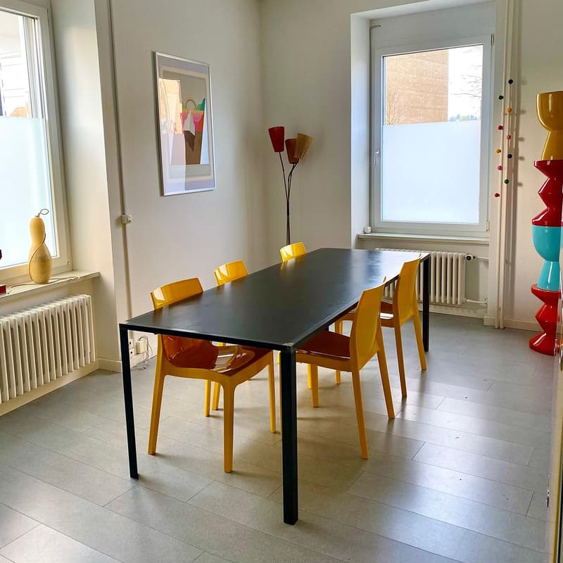 Dining table with black top and yellow chairs, artwork on the wall, decorative vase, and a window with a view of the outdoors.