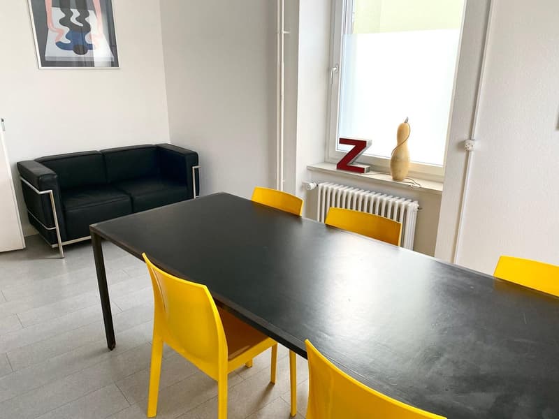 Black leather sofa, black rectangular dining table, yellow chairs, artwork on the wall, radiator, and a decorative object on the windowsill