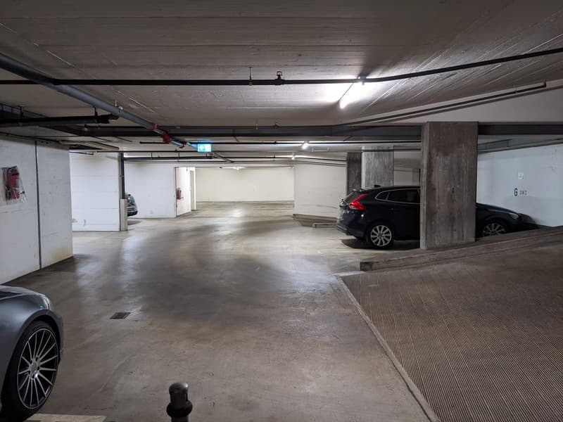 underground parking lot, concrete floor, white walls, two cars parked, one car in the ramp