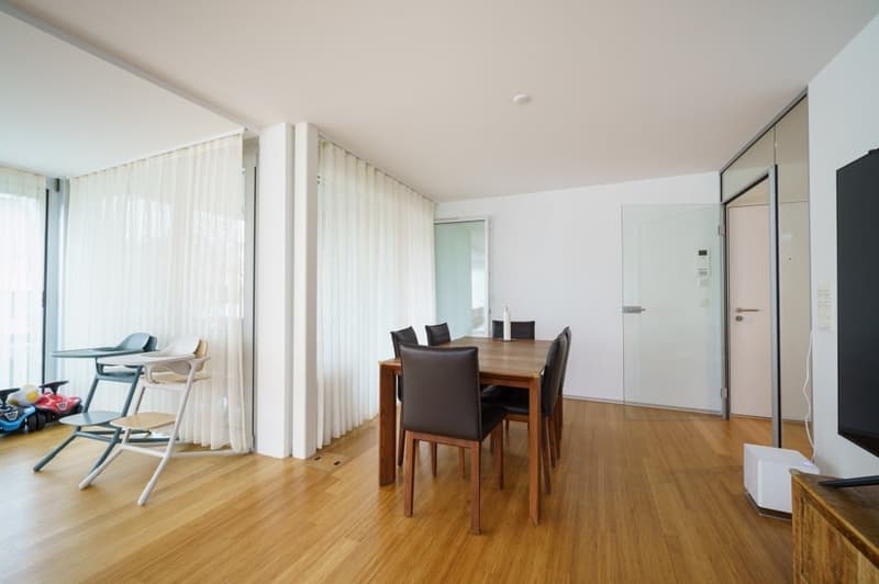 Dining room with wooden floor, glass doors, white walls, dining table, chairs, white curtains, and a television.