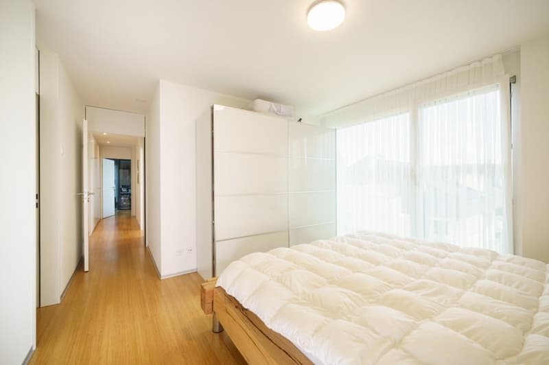 Bedroom with wooden floor, white walls, large windows, bed with white duvet, white wardrobe, ceiling light