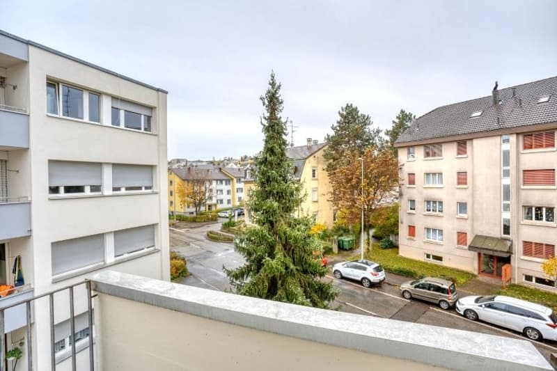 A balcony view of a building with cars parked in front, a tree, and other buildings in the distance.