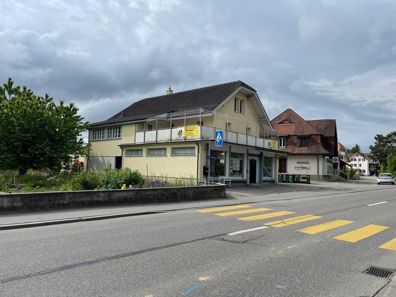 A two-story commercial building with a yellow exterior, a tiled roof, and several windows. The building has a grocery store called 'Swissco' on the ground floor. There is a crosswalk in front of the building and the surrounding area appears to be a reside