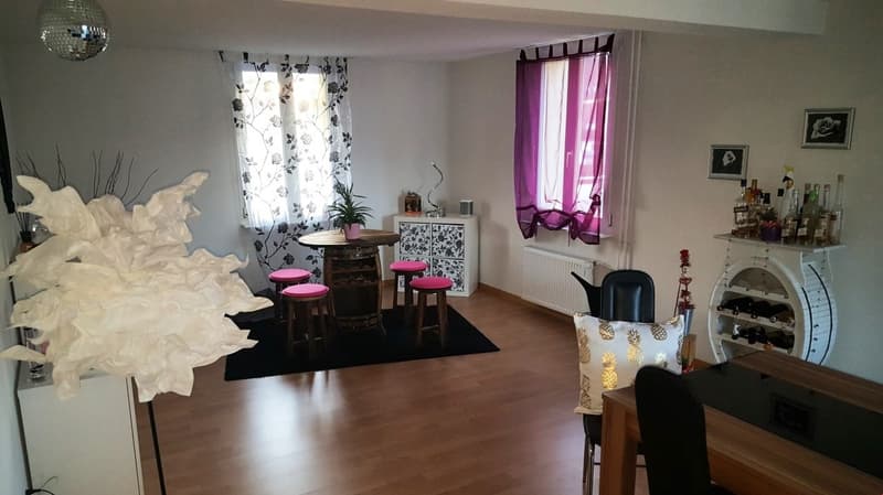 Spacious living room with hardwood floors, white walls, large windows, floral curtains, a disco ball, a wooden table with pink stools, a cabinet with decorative items, and a fireplace. The room also has a bar area with liquor bottles and a wine rack.