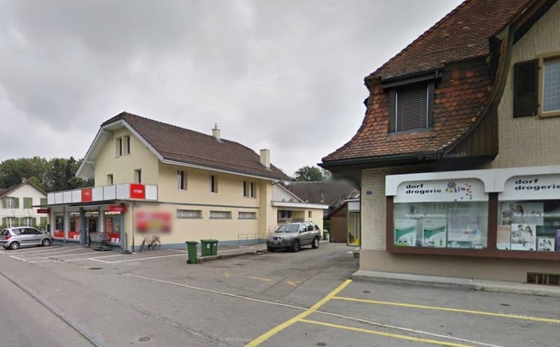 Two-story commercial building with a grocery store and other shops, parking spaces in front, residential buildings in the background, cloudy sky