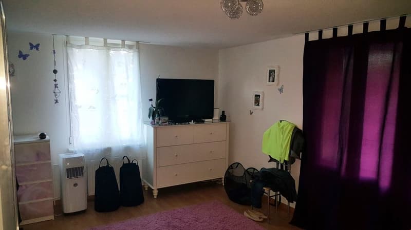 Bedroom with white walls, hardwood floor, dresser, TV, and purple curtains. There is a balcony or terrace visible and a private washing machine in the room.