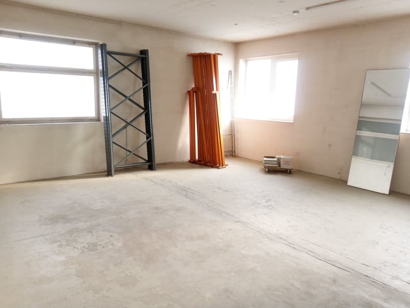 Empty room with white walls, concrete floor, two windows, metal rack, orange poles, mirror, and a trolley with boxes.