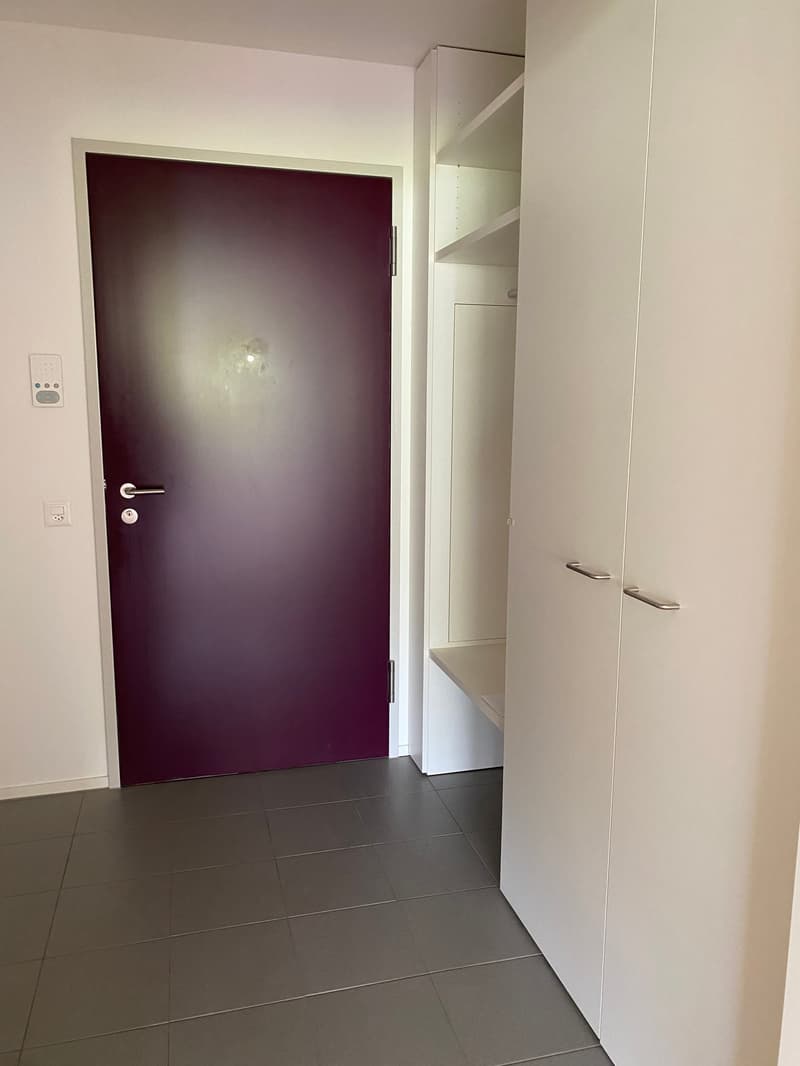 An empty room with a door and a shelf, tile flooring, white walls, and a closed door with a handle and lock