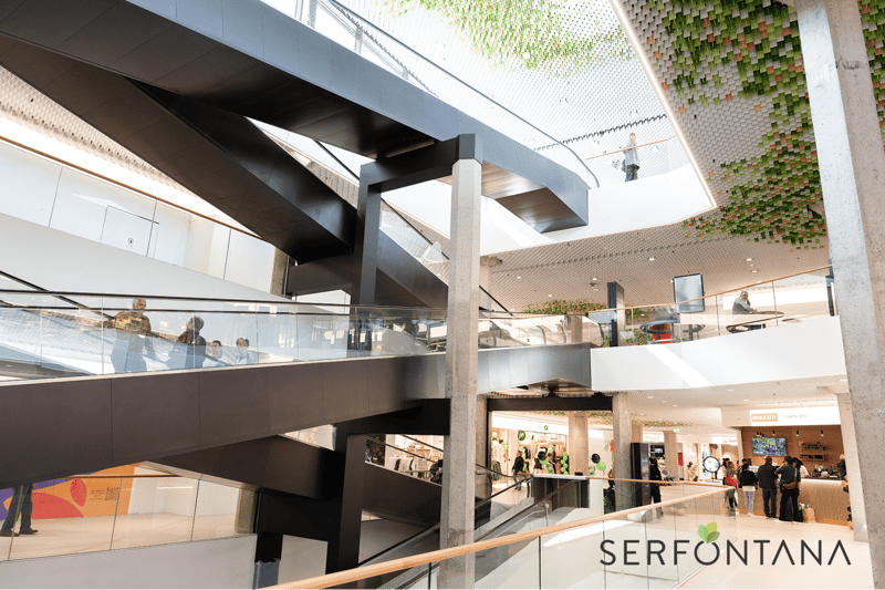 modern building, glass walls, escalators, multiple levels, people walking, shops, green plants