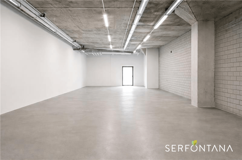 A large, empty room with concrete flooring and white walls. A doorway is located in the center of the room. Overhead lighting is affixed to the ceiling, and an entrance with a glass door is situated on the left side of the image.