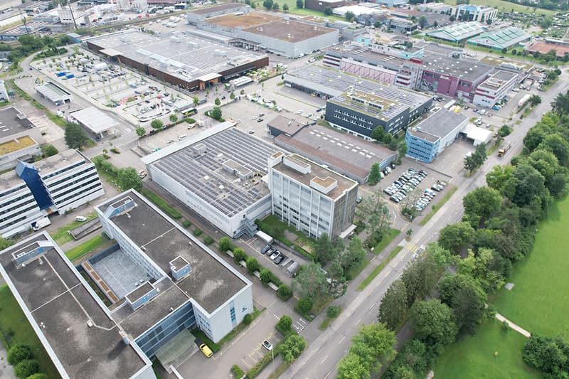 Industrial complex with multiple buildings, surrounded by parking lots, trees, and open green spaces.