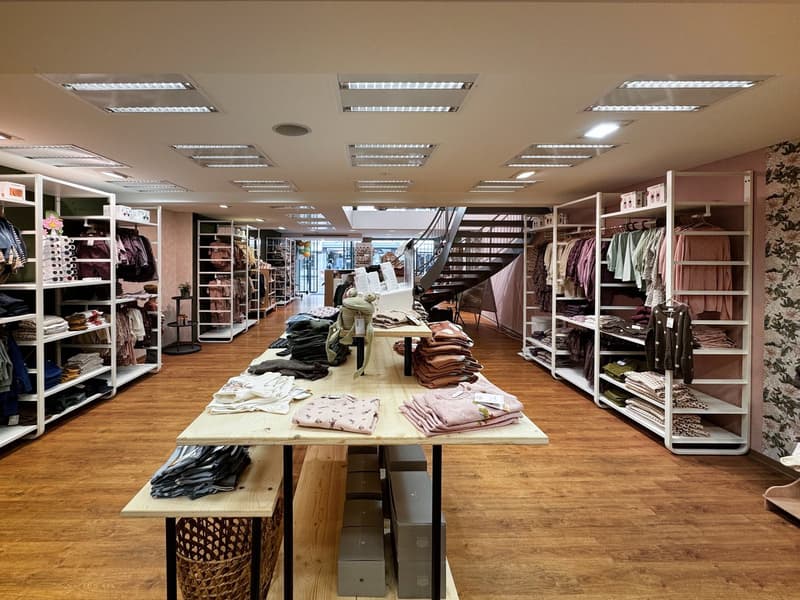 An indoor space resembling a boutique with clothing items on racks, long wooden tables with merchandise, wooden flooring, and a spiral staircase leading to another level. The walls are adorned with floral wallpaper and shelves are filled with various item