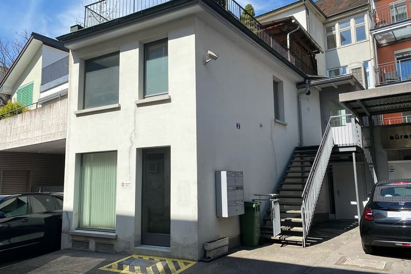 2-story building with a modern, minimalist design. It has large windows, a balcony, and a parking space in front of the building.