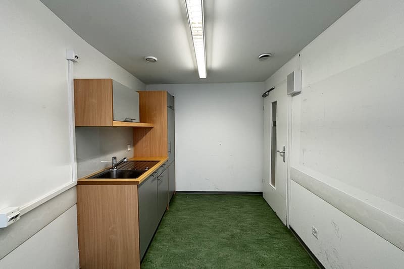 The image shows a small studio apartment with a kitchenette area. The kitchenette has wooden cabinets and a sink. The floor is covered with green carpet. The room has a white wall and a ceiling with a long light fixture.