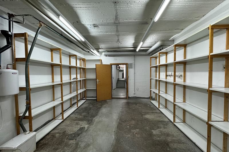 The image shows a large storage room or warehouse space with multiple rows of shelving units along the walls. The shelves appear to be made of wood and metal, and the room has a concrete floor and a high ceiling with exposed pipes and lighting fixtures. T