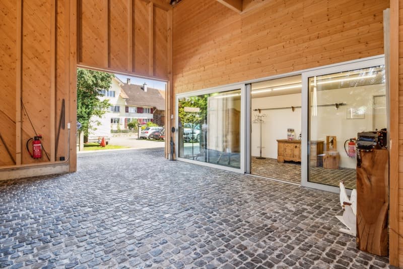 Open garage with glass doors, wooden walls, cobblestone flooring, fire extinguishers on the wall, and a view of a house and car parked outside