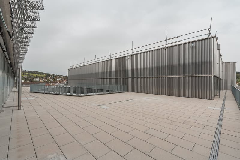 Large industrial-style building with a corrugated metal exterior, surrounded by a paved plaza with a tiled floor. The building appears to be a modern architectural structure with a minimalist design.
