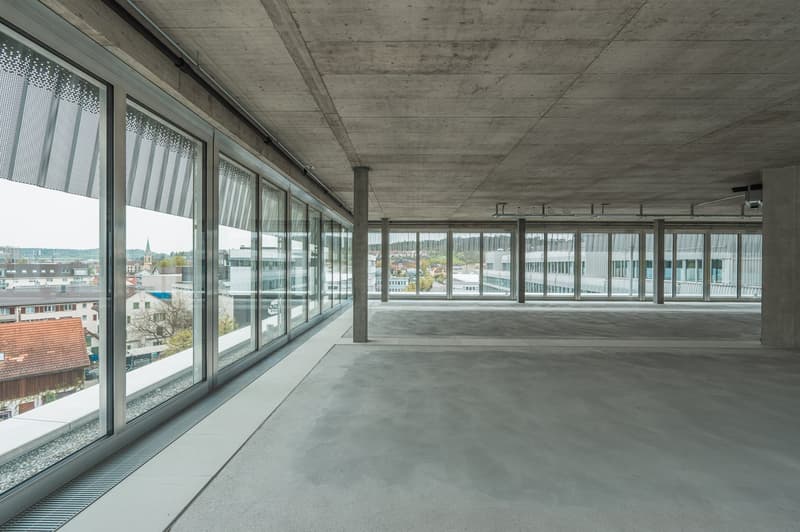 Large open-plan office space with concrete ceiling and floor, floor-to-ceiling windows offering a view of the city skyline