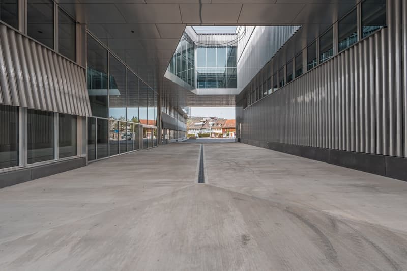 The image shows a modern, multi-story commercial building with a glass facade and a long, open walkway in the center. The building has a sleek, industrial design with metal panels and a large skylight overhead. The walkway is made of concrete and leads to