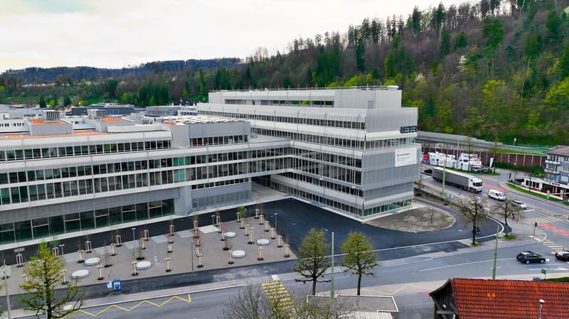 A large, multi-story office building with a modern, glass and steel design. The building has a plaza in front with various seating areas and landscaping. The surrounding area includes forested hills and other commercial buildings.