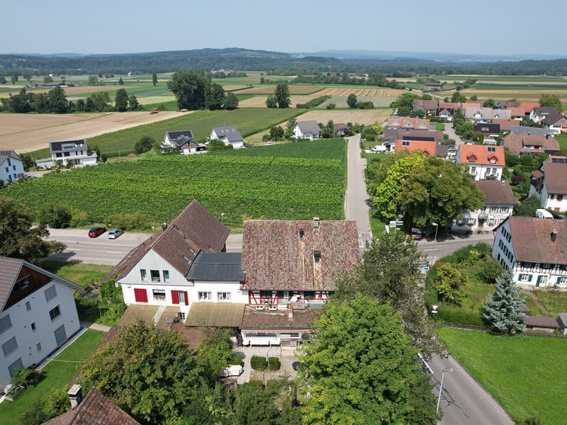 House located in countryside, with surrounding fields, raised ground floor, garage, solar panels, green areas, multiple windows, neighboring houses, cars on street