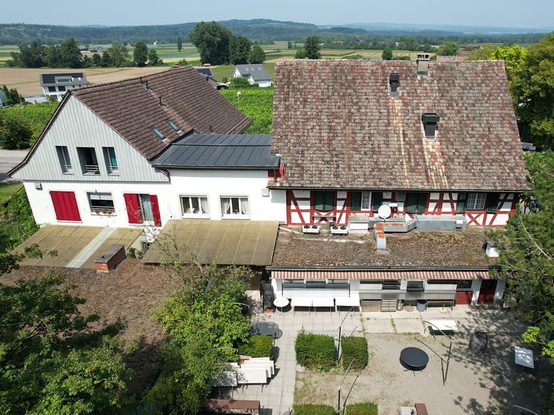 Aerial view of a two-story house with a tiled roof, brick walls, red shutters, multiple windows, a covered outdoor dining area, and an attached garage. The house is surrounded by greenery and offers a scenic view of the countryside.