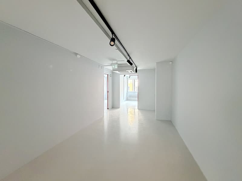 A long, narrow room with white walls and a white floor. There are two doors on either side and two track lights on the ceiling.