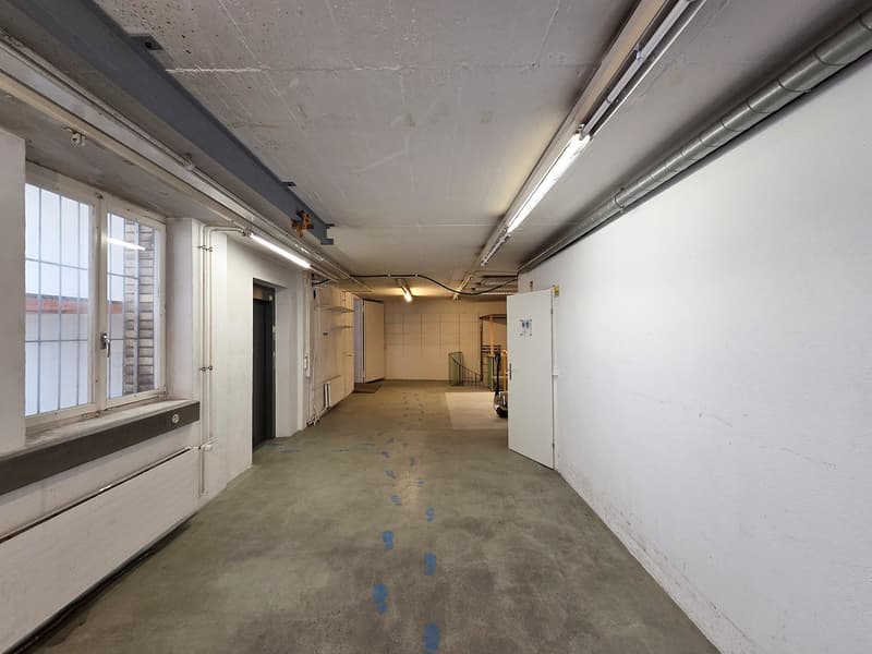 Empty basement with gray concrete floor, two open doors, large window, white walls, radiator, pipes, light fixtures, and footprints on the floor