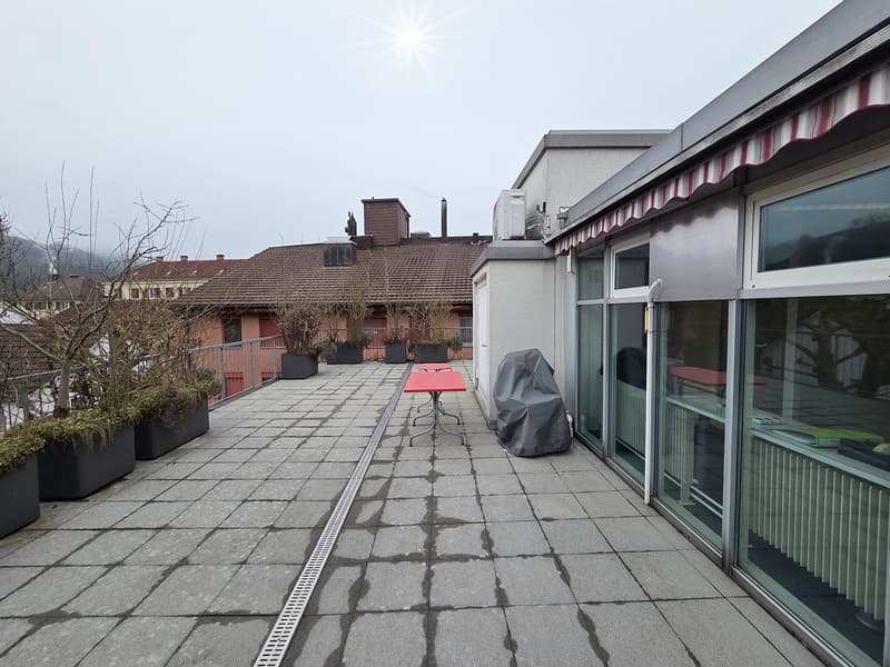 Terrace with tiled floor, metal railing, red table and chairs, covered with red and white awning, with plants in large pots, and overlooking another house.