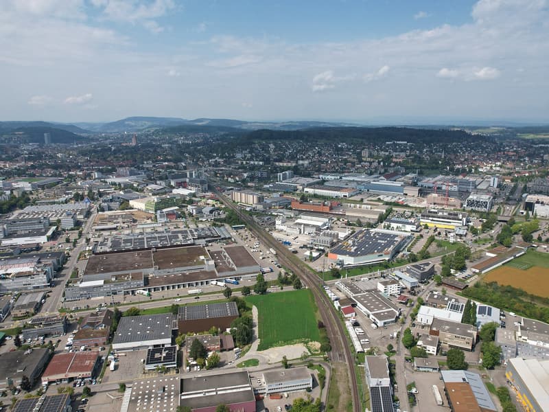 Aerial view of a city with many buildings, roads, and greenery. Several large industrial buildings are visible, along with smaller residential and commercial structures. The area is well-developed with a mix of urban and suburban features.