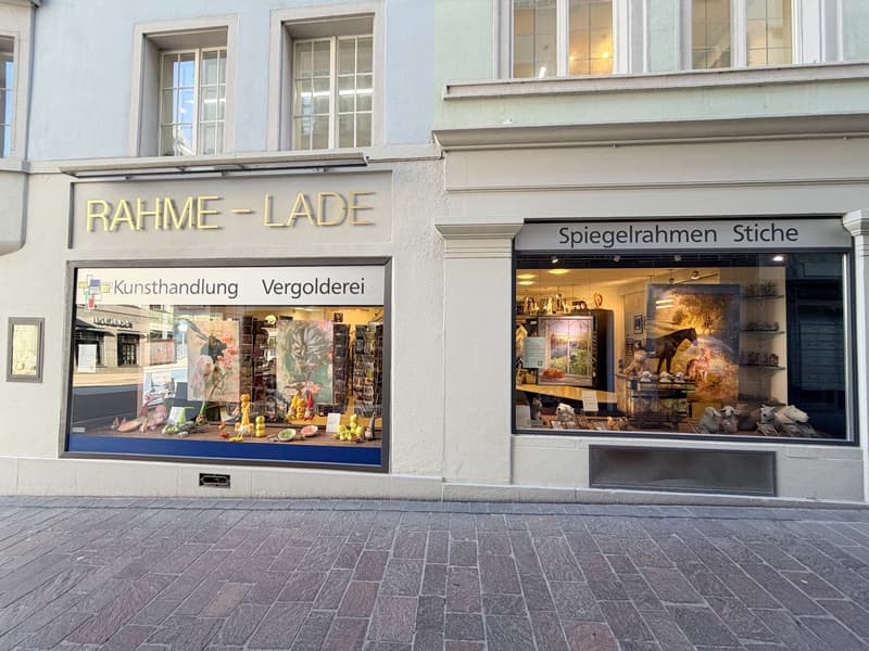 Facade of a building with two shops, one named 'RAHME-LADE' and the other 'Spiegelrahmen Stiche', both with glass windows displaying items inside.