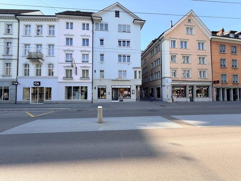 A street with several buildings, one of which has a sign that reads 'RAHME-LADE'. The buildings have multiple windows and some have balconies. The street is empty, and there is a person walking on the sidewalk.