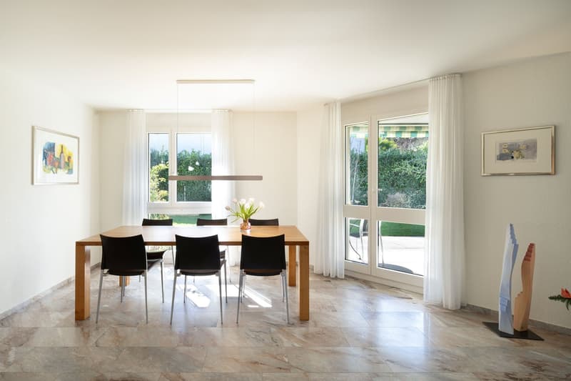 Dining room with wooden table, black chairs, white walls, large windows, white curtains, flower vase, artwork, marble floor