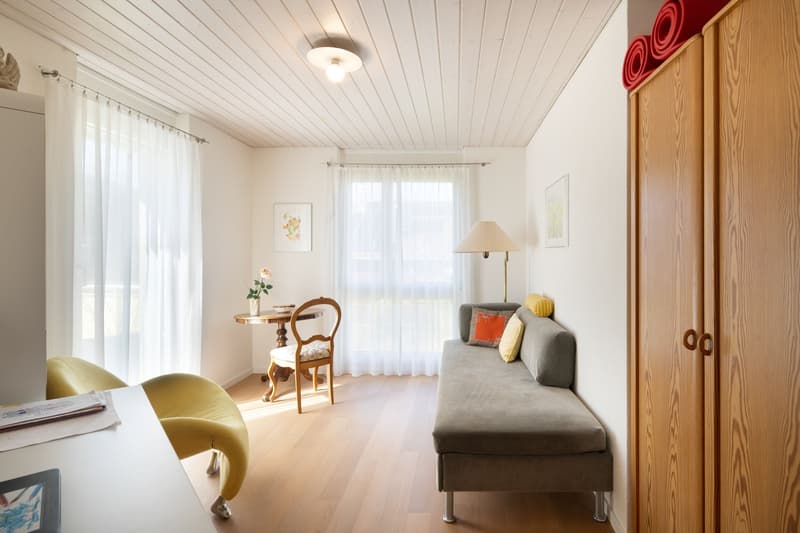 A room with a wooden floor, white walls, a gray sofa, a wooden chair, a small table, a lamp, a wooden cabinet, and a white curtain.