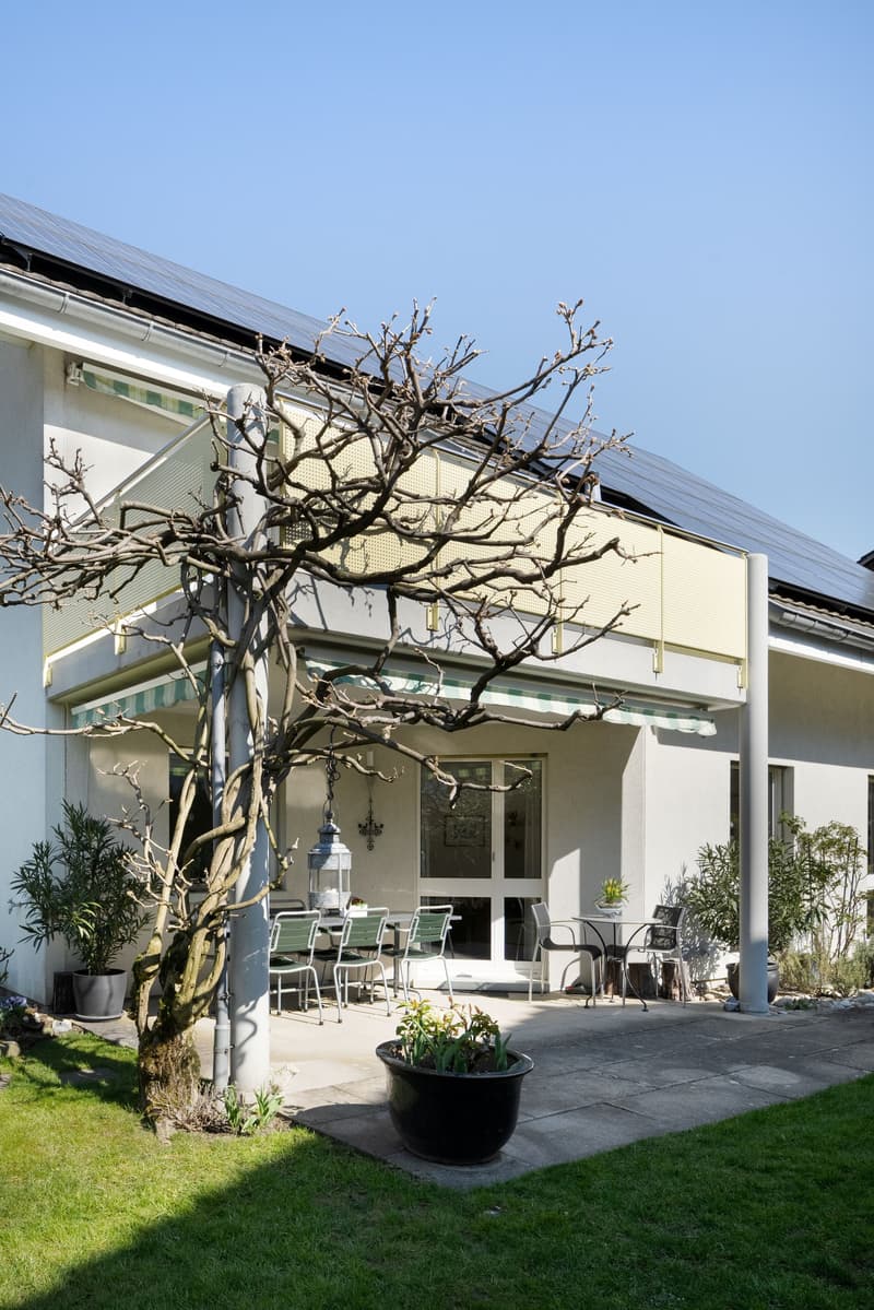 white house, solar panels on the roof, balcony with awning, outdoor dining area, potted plants