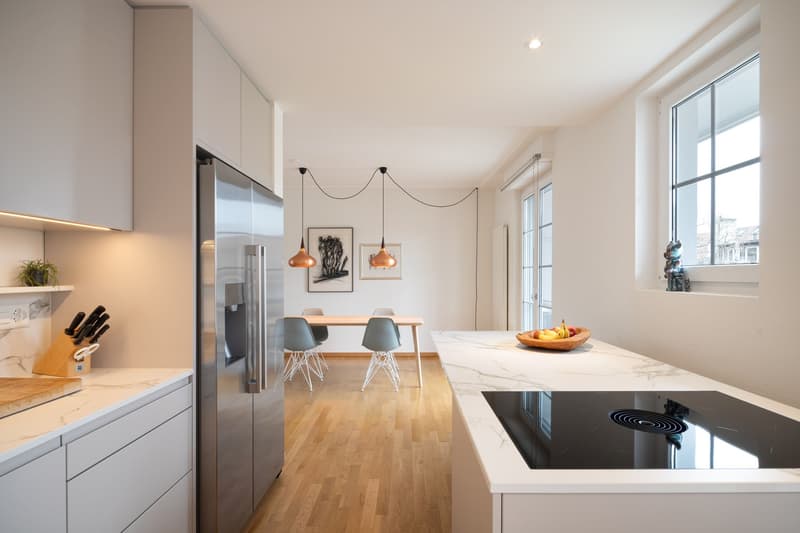 modern kitchen with marble countertops, stainless steel refrigerator, induction cooktop, wooden floor, dining table with chairs, pendant lights, artwork on the wall, window with view