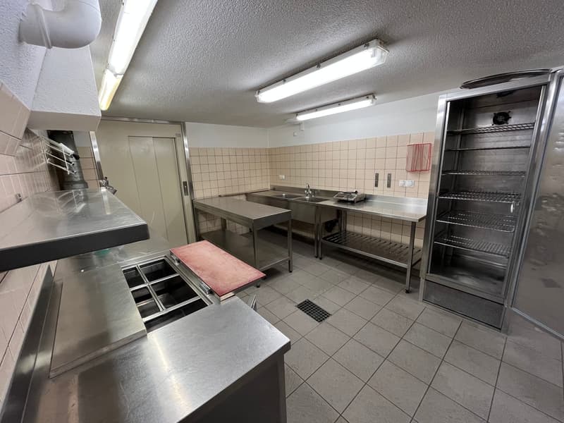 Commercial-grade kitchen with stainless steel countertops, sinks, and appliances, including a dishwasher and washing machine. The kitchen is tiled and has fluorescent lighting overhead.