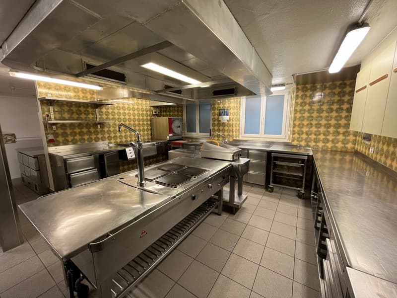 Large commercial kitchen with stainless steel appliances and equipment, including a stove, oven, and dishwasher. The kitchen has tiled floors and walls with a yellow and green tile pattern. There are multiple workstations and storage areas throughout the