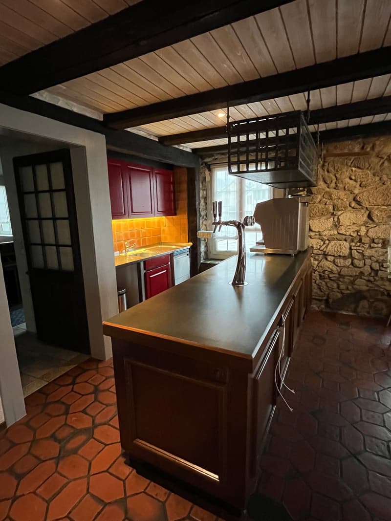 Rustic kitchen with exposed wooden beams, red cabinets, orange tile backsplash, stone walls, and a kitchen island with a dark countertop. There is a fireplace visible in the room.