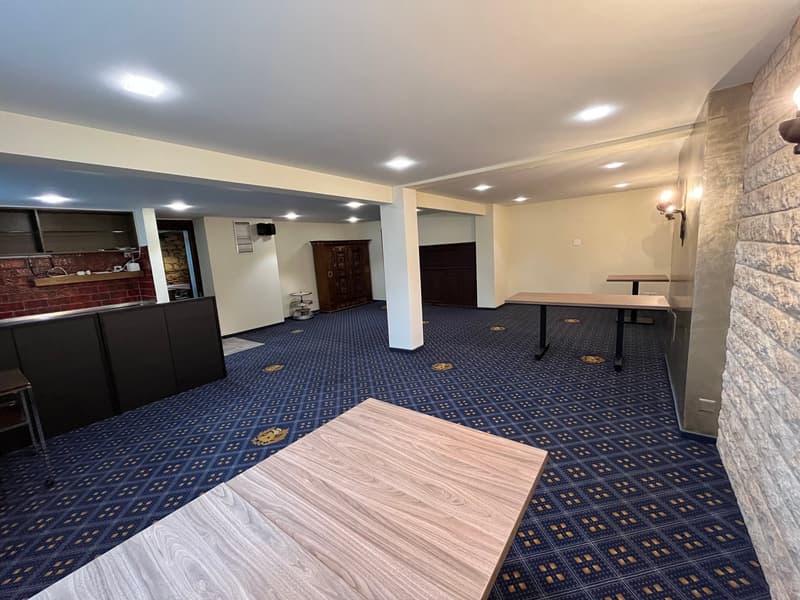 Spacious basement with a kitchenette area, dining tables, and a patterned blue and white carpet flooring. The room has recessed lighting and a brick accent wall.