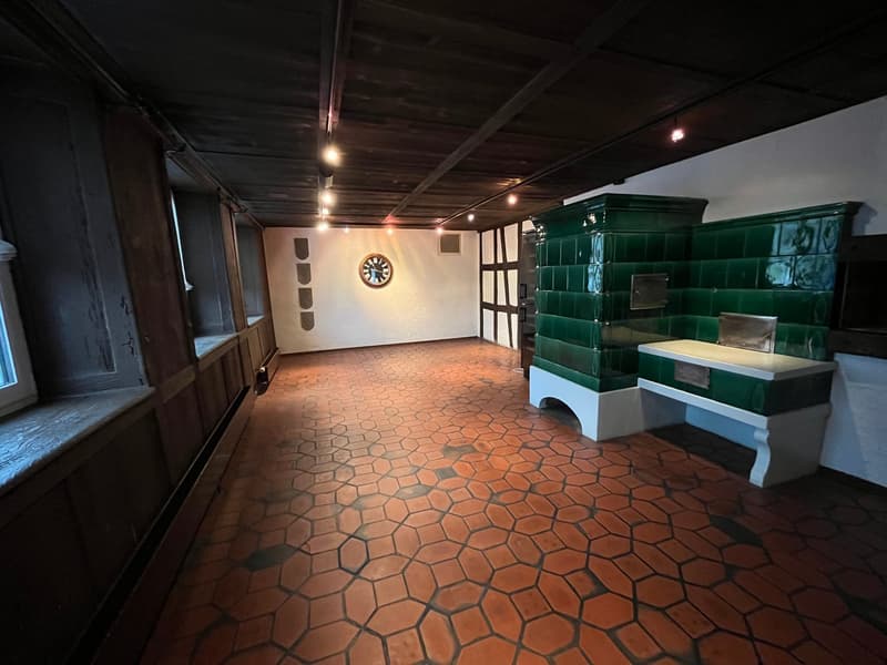This image shows a room with a tiled hexagonal floor, exposed wooden ceiling beams, and a large green tiled stove or fireplace against the wall. The room has a rustic, traditional feel with the wooden and tile elements. There is also a clock on the wall a