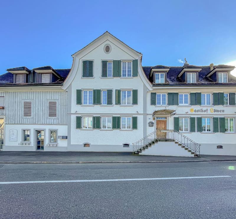 3-story building with white exterior, green shutters, and a gabled roof. The building has a balcony on the second floor and a set of stairs leading up to the main entrance. The building appears to be a hotel or inn, as indicated by the sign 'Gasthof Loewe