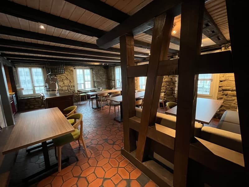 Rustic dining room with exposed wooden beams, stone walls, hexagonal tile flooring, wooden tables and chairs, and a fireplace visible.