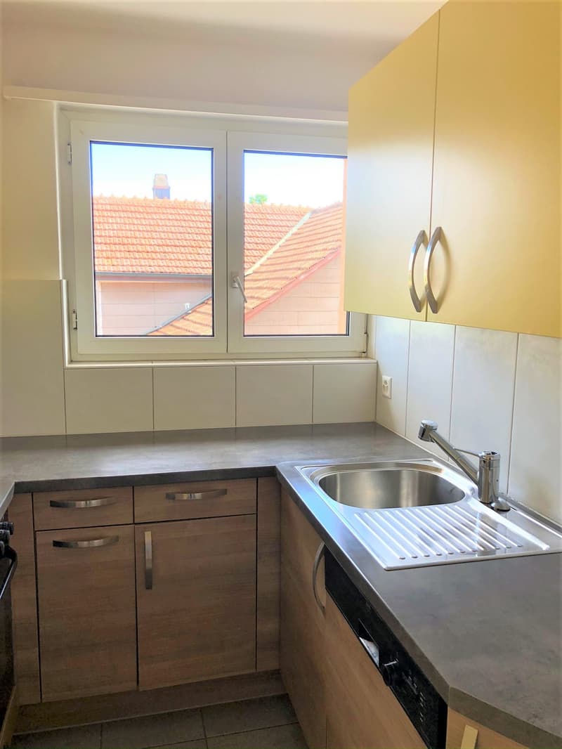 A small kitchen with a sink, faucet, a drain board, an oven underneath the sink, four wooden cabinets, and a window.