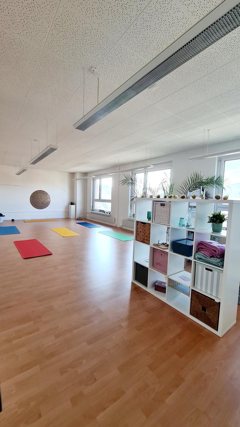 Yoga studio with wooden floors, suspended lamps, yoga mats, white shelves, and potted plants. The room has a minimalist design, with a focus on providing a peaceful and comfortable space for yoga practice.