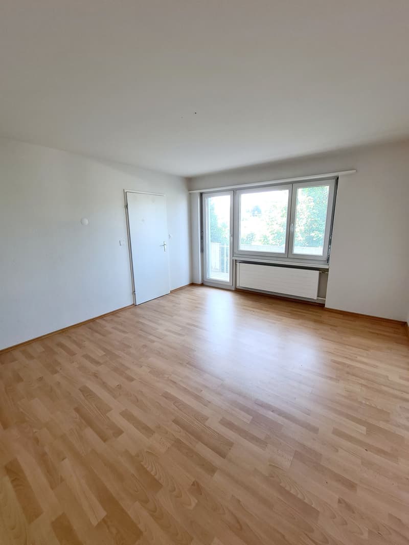 Spacious room with hardwood flooring, large windows providing natural light, and a white wall. The room appears to be empty, ready for furnishing and decorating.