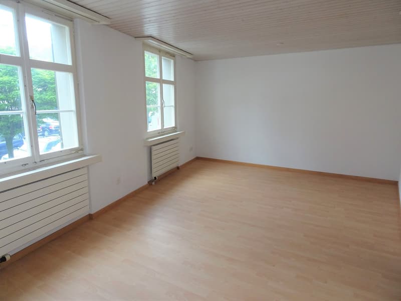 This image shows a spacious room with white walls, a wooden floor, and large windows that provide natural light. The room appears to be empty, with a radiator installed along one of the walls.