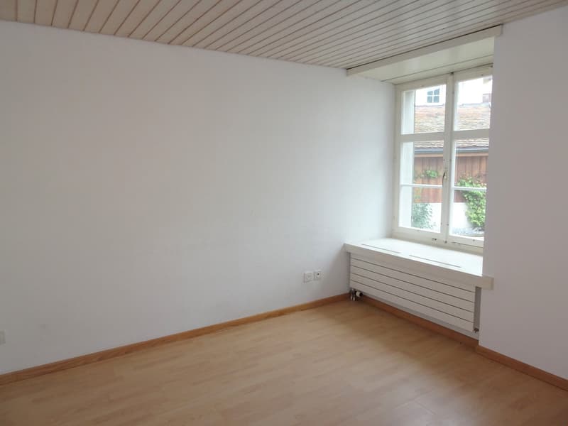 This image shows a simple, minimalist room with white walls, a wooden ceiling, and a large window overlooking a garden or outdoor area. The room has a hardwood floor and a radiator or heating unit installed along the wall. The room appears to be empty, wi